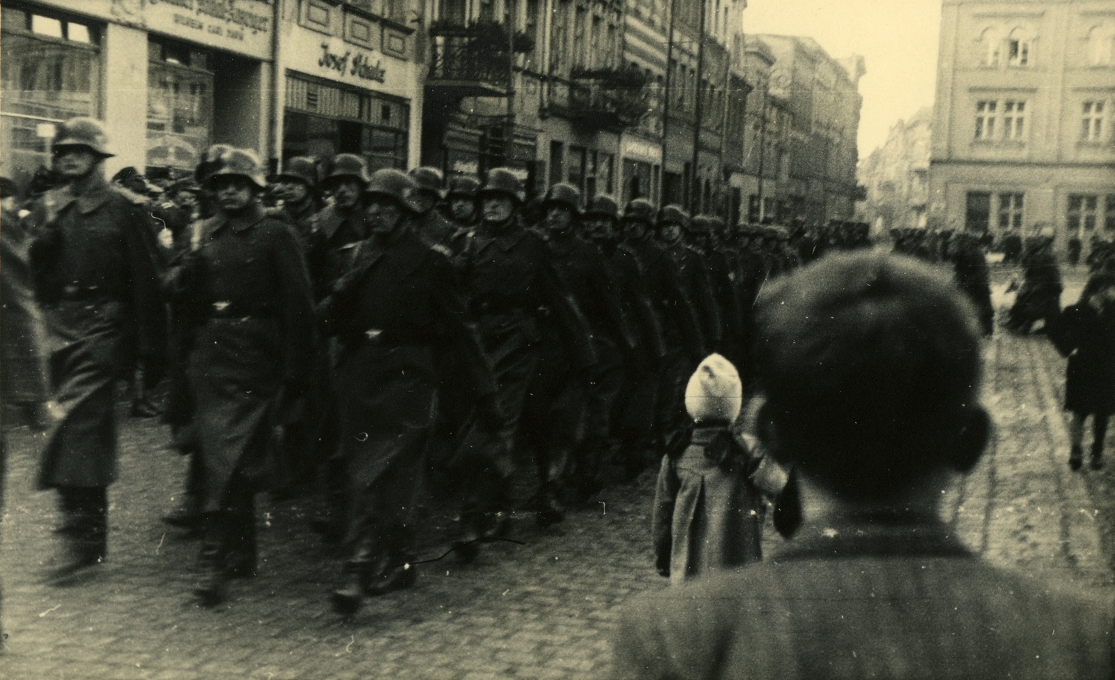 Fotografia zbiorowa. Na rynek w Lesznie wkraczają maszerujące oddziały niemieckiego wehrmachtu. Żołnierze ubrani są w wysokie buty i zimowe płaszcze spięte pasami. Na głowach mają hełmy, na ramionach karabiny. Paradzie przyglądają się stojący na chodnikach przechodnie. W tle widać fasady przyrynkowych kamienic i budynki przy obecnej ulicy Słowiańskiej.