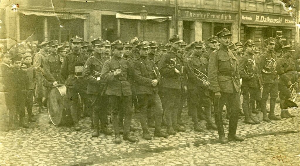 Fotografia zbiorowa w zielonkawym odcieniu. Członkowie orkiestry wojskowej stoją w oczekiwaniu na rozpoczęcie uroczystości. Pod pachami trzymają trąbki i waltornie. Na czele grupy stoi kapelmistrz z batutą w prawej ręce. W głębi żołnierz przytrzymujący duży bęben. Obok niego widać grupę chłopców w mundurkach szkolnych. W tle przyrynkowe kamienice z reklamami w języku niemieckim i polskim: Theodor Freundlich i Hurtowania A. Dzikowski. 