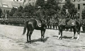 Fotografia zbiorowa. Na placu koszarowym stoją w dwóch rzędach żołnierze w pełnym rynsztunku. Karabiny z nałożonymi bagnetami przytrzymują na wysokości piersi. Z przodu dowódca 17 Pułku Ułanów pułkownik Aleksander Pragłowski składa raport generałowi brygady Sergiuszowi Zahorskiemu. Oficerowie siedzą na koniach. Z lewej strony widoczny poczet sztandarowy. W głębi żołnierze na koniach z uniesionymi do góry lancami, na których powiewają proporczyki. W tle wysokie budynki koszarowe.