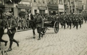 Na fotografii kolumna żołnierzy leszczyńskiego garnizonu podczas defilady. Wszyscy w mundurach nowego wzoru, wprowadzonych na stan w latach 1935-1937. Na nogach mają buty wojskowe i owijacze, na głowach stalowe hełmy. Karabiny zawieszone na piersiach. Do plecaków przytroczone koce. W środku jeden z żołnierzy prowadzi konia z taczanką, na której zamontowano karabin maszynowy. Na chodniku stoją mieszkańcy – dorośli i dzieci oraz odbierający defiladę oficerowie na trybunie. W tle przyrynkowe kamienice z reklamami w języku polskim. Widoczne są szyldy kilku sklepów: bonboniera, Fabryczny skład i Józef Danielak. Na budynkach wiszą flagi narodowe.