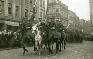 Fotografia przedstawia grupę ułanów wjeżdżających na leszczyński rynek. W pierwszym rzędzie poczet sztandarowy. Żołnierz w środku, trzymający pułkowy sztandar, siedzi na białym koniu. Na piersi ma ozdobną szarfę. Ułani po bokach trzymają uniesione szable. Żołnierz w drugim rzędzie ma zawieszony na plecach karabin. Kolejni trzymają lance z proporczykami. Na chodniku tłum obserwujących wydarzenie mieszkańców. Z lewej strony grupa mężczyzn w grubych płaszczach. W tle kamienice przy rynku i ulicy Kościańskiej (obecnie Gabriela Narutowicza).