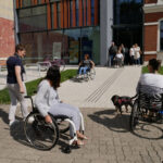 Kilkanaście osób znajduje się na świeżym powietrzu przed nowoczesnym budynkiem. Część z nich porusza się na wózku inwalidzkim. Zdjęcie przedstawia szkolenie w ramach programu "Kultura dostępna" dotyczące potrzeb osób z niepełnosprawnością ruchu.