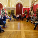 Sala muzealna, warsztaty dla pracowników muzeum na temat dostępności dla osób z niepełnosprawnością słuchu. Uczestnicy siedzą na krzesłach. Pomieszczenie zdobią obrazy postaci historycznych.